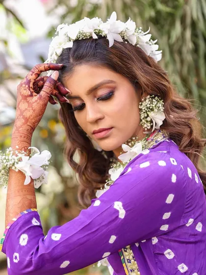 A vibrant and fresh look for a Phool Haldi ceremony. The makeup features warm, glowing skin and defined eyes, designed to harmonize with the purple bandhani outfit and the beautiful fresh flower jewelry.