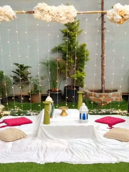 A simple and chic boho picnic arrangement in a garden. The setup features a low table with floor cushions, framed by a backdrop of white drapes, pampas grass, and delicate floral accents.