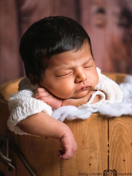 A close-up portrait of a newborn posed comfortably in a wooden bucket against a wood-paneled backdrop.