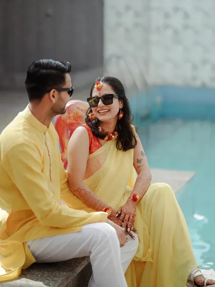 A candid shot of a couple sharing a laugh by the poolside during their Haldi ceremony, both dressed in cheerful yellow outfits.