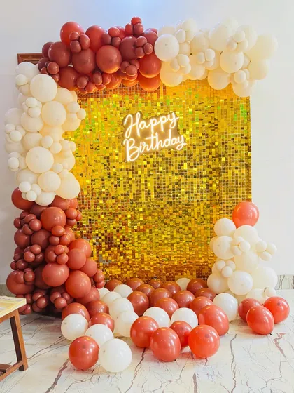 A chic birthday backdrop with a gold shimmer wall, a 'Happy Birthday' neon sign, and an organic balloon arch in rustic red and cream colors.