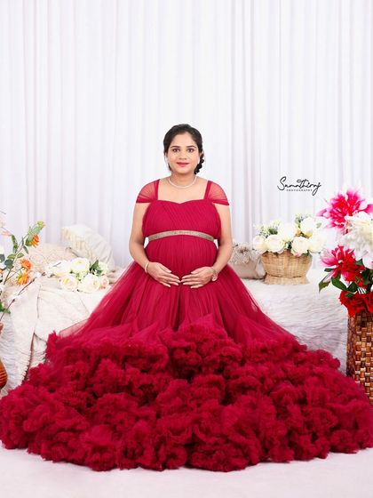 A full-length portrait of a mother-to-be in a stunning red ruffled gown. The simple floral studio setup allows the dress and her radiant glow to take center stage.