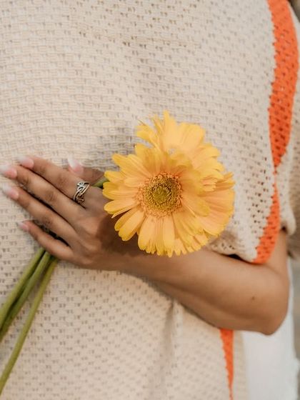 A creative and sweet detail shot. The bride-to-be holds a yellow flower behind her back, adding a touch of playful romance to their picnic-themed shoot.