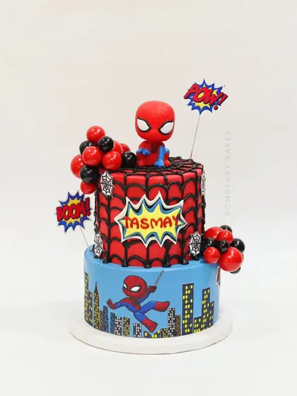 A two-tier Spiderman cake that travelled all the way to Alibag. The design includes a city skyline, web details, and a Spiderman bobblehead topper.