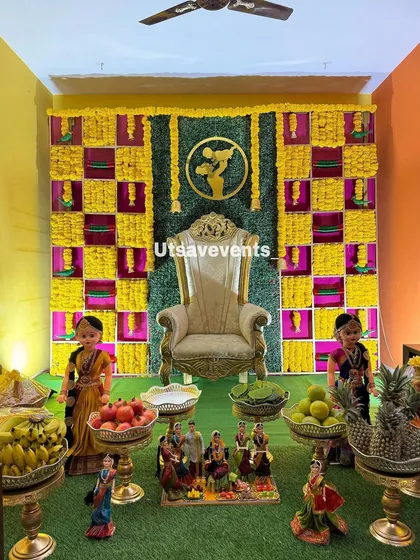 A closer look at a home baby shower setup, showing the detail of the marigold checkered backdrop, the golden throne, and the arrangement of traditional dolls and fruits.