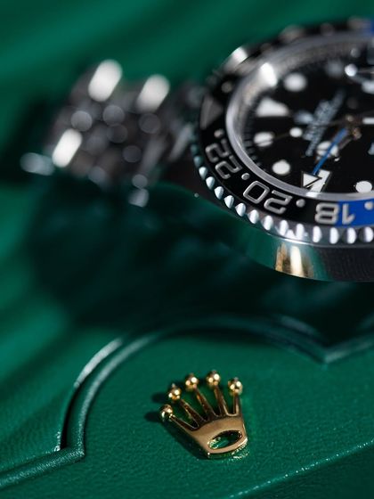 The iconic Rolex crown emblem, detached from the box clasp, sits beside the watch. This shot emphasizes the branding and luxury status of the pieces we handle.