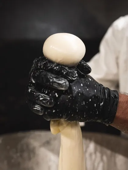 A chef's gloved hand expertly forming a ball of fresh mozzarella. The specks on the glove are from the whey, a natural part of the process.