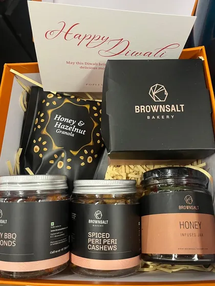 This is an unboxed view of a custom corporate hamper for Popley Eternal. It includes our Honey & Hazelnut Granola, Spicy BBQ Almonds, Spiced Peri-Peri Cashews, and a Honey Infused Jar, all accompanied by a personalized Diwali card.
