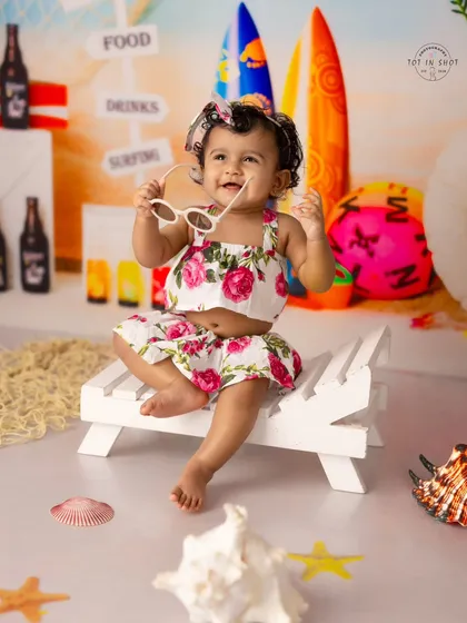A happy, playful moment captured during a beach-themed sitter session. The floral outfit and stylish sunglasses add a touch of personality to this sunny portrait.