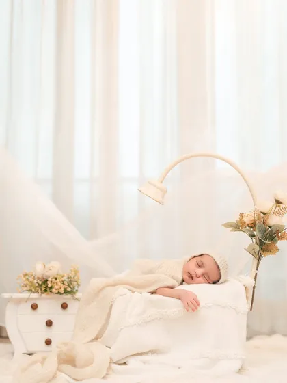 A vertical composition focusing on the sleeping baby, wrapped snugly in a basket. The flowing white curtains in the background add a soft, dreamy quality to this classic baby portrait.