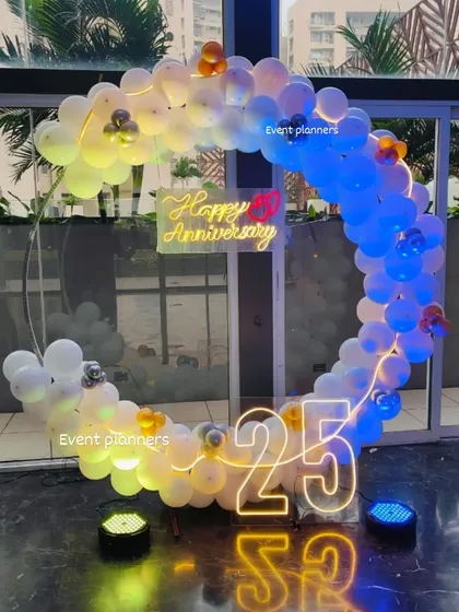 Celebrate your milestones in style. This 25th-anniversary decoration features a modern semi-circular balloon arch with multi-color mood lighting, a custom 'Happy Anniversary' neon sign, and large marquee numbers, creating an elegant and festive atmosphere.