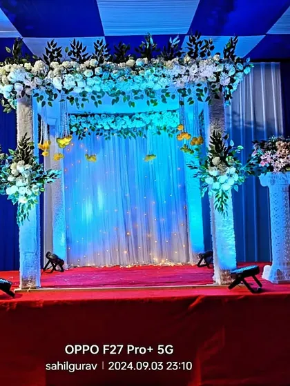 A mandap-style decoration for a stage. The structure is adorned with white flowers and greenery, with blue and white drapes creating a serene and elegant setting.