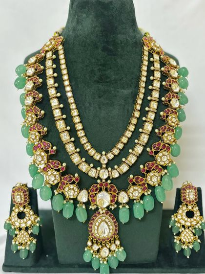 This layered bridal set features a choker and a long necklace with green beads and red Meenakari work. It includes matching earrings for a complete rental package.