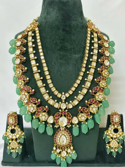 This layered bridal set features a choker and a long necklace with green beads and red Meenakari work. It includes matching earrings for a complete rental package.
