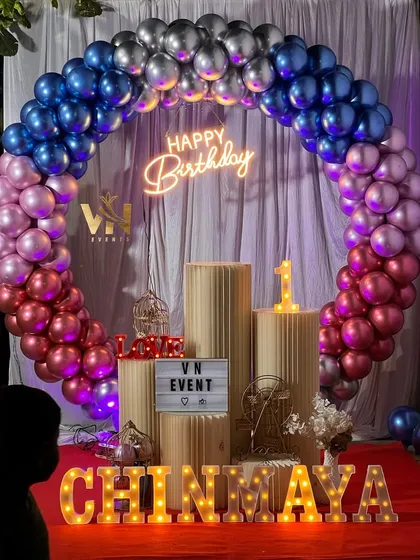 A full view of a birthday setup with a colorful chrome balloon arch in blue, silver, pink, and red. The lit-up name and neon sign make it perfect for photos.