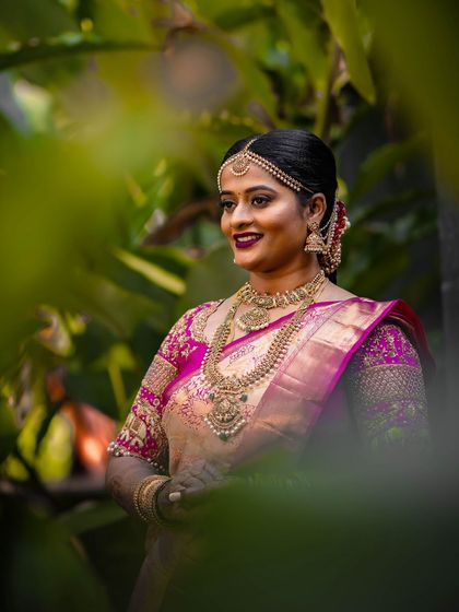 A beautiful portrait of the bride in her peach and pink saree, surrounded by lush greenery.