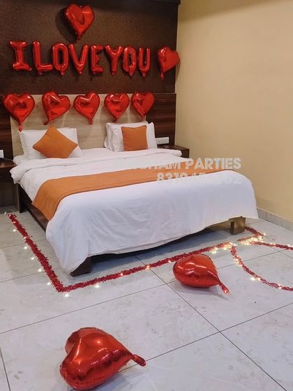 A hotel room decorated for a romantic occasion with "I LOVE YOU" foil balloons and a heart made of rose petals on the floor.