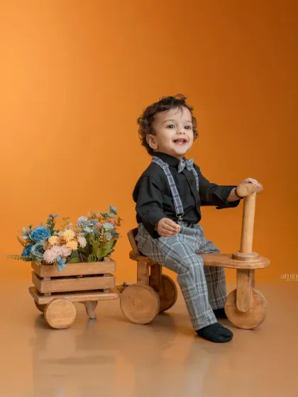 A dapper little boy in plaid pants and a bow tie enjoys a ride on a wooden tricycle. The attached cart full of flowers adds a whimsical touch to this charming studio portrait.