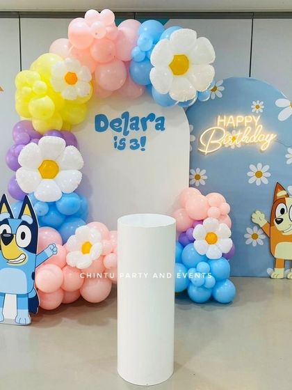 A fun and colorful Bluey-themed birthday decoration for a third birthday. The setup includes character cutouts of Bluey and Bingo, daisy-shaped balloons, and a 'Happy Birthday' neon sign.
