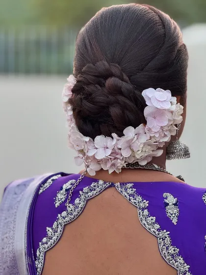 An elegant braided updo for Dr. Indhu's reception. The bun is encircled with delicate pink flowers, creating a soft and romantic look that complements her silver jewellery.