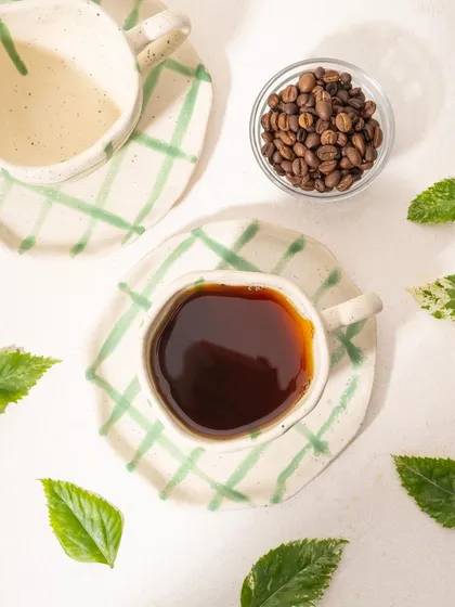 A flat lay of a Homesmith coffee cup, styled with fresh leaves and coffee beans. The strong shadows from the direct light create a modern, graphic look.