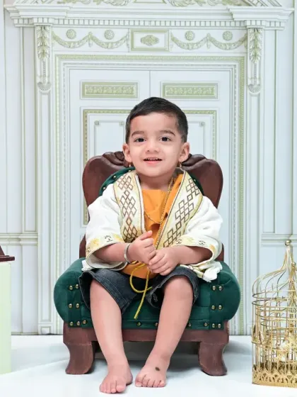 This little boy smiles warmly for his vintage-style portrait. Seated in a green armchair, the scene is set with classic props that evoke a sense of old-world elegance.