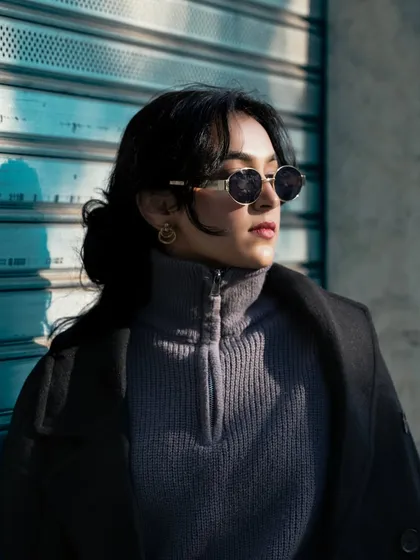 A close-up portrait of a model with sunglasses against a blue shutter. The hard sunlight creates interesting shadows, adding a high-fashion, editorial feel to the image.