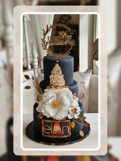 An elegant three-tier corporate cake in black and gold, featuring company logos and beautiful white sugar flowers.