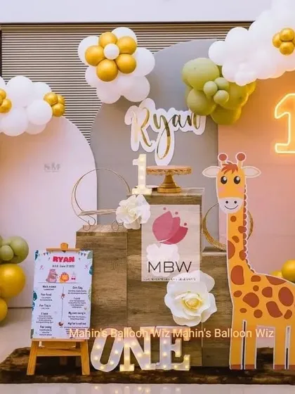 A cheerful and modern first birthday setup with a friendly giraffe character. The design uses a warm color palette of olive green, orange, and gold, complemented by a neon "1" and a personalized milestone board.