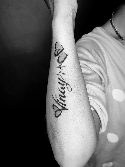 A black and white shot of the "Vinay" name tattoo, emphasizing the clean script and the sharp line of the heartbeat.