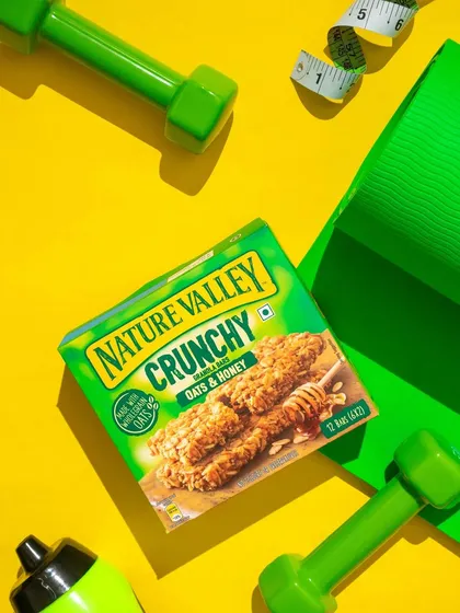 A high energy flatlay for a healthy snack bar, surrounded by workout essentials like a yoga mat, dumbbells, and a measuring tape. The bright yellow background and green props create a vibrant, fitness focused composition.