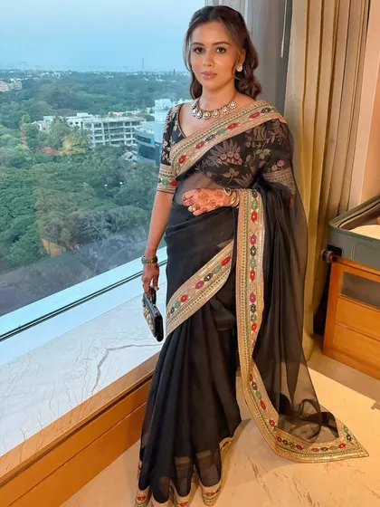 The client poses by a window, the natural light highlighting her glowing makeup and the saree's intricate border.