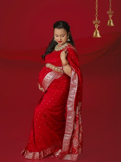 A stunning portrait of a mother-to-be in a vibrant red Banarasi saree. The traditional temple bells and rich red background create a divine and celebratory feel for this ethnic maternity shoot.