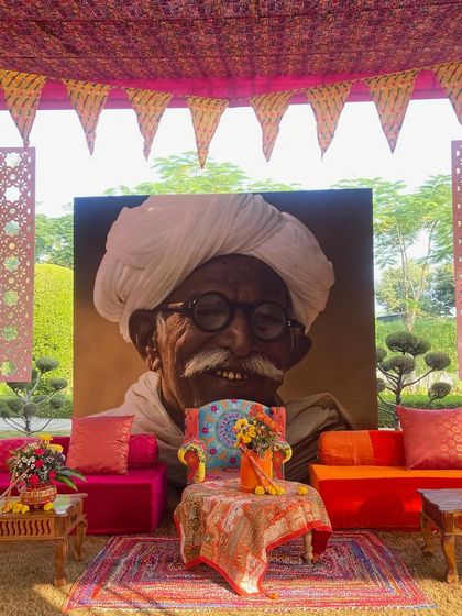 A unique photo point at the Haldi ceremony, featuring a large portrait of a man in a traditional turban. The colorful, low seating in front creates a perfect spot for guests to take memorable pictures.
