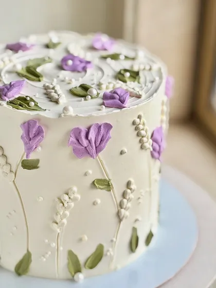 A delicate floral cake featuring hand-piped purple flowers and greenery on a white buttercream base. Tiny edible pearls add a finishing touch of elegance.