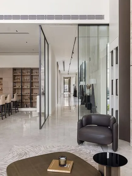 This open-plan living area in the Ananda residence connects the lounge, kitchen, and a long corridor. The design uses glass partitions and a consistent marble floor to create a feeling of endless space and light.