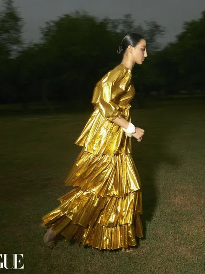 An editorial for Vogue that captures movement and drama. The styling features a tiered, metallic gold dress that creates a fluid, sculptural shape as the model moves, blending high fashion with a sense of dynamic energy.