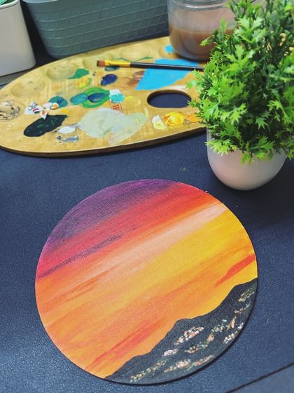 A finished sunset painting on a round canvas, placed next to a wooden paint palette.
