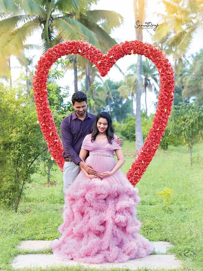 A romantic outdoor portrait of a couple framed by a heart-shaped floral prop. The soft pink gown and the lush greenery create a perfect fairytale setting.