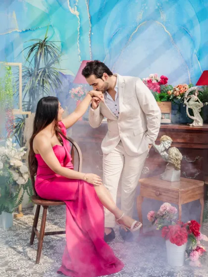 A chivalrous moment captured amidst a whimsical, artistic set. The groom kisses the bride's hand, surrounded by flowers and a dreamy, smoke-filled atmosphere.