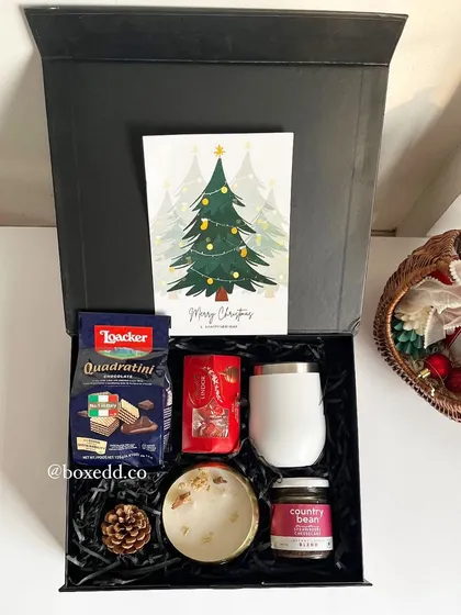 A festive Christmas hamper in a black box, featuring a "Merry Christmas" card, gourmet wafers, a travel tumbler, and other holiday-themed treats.