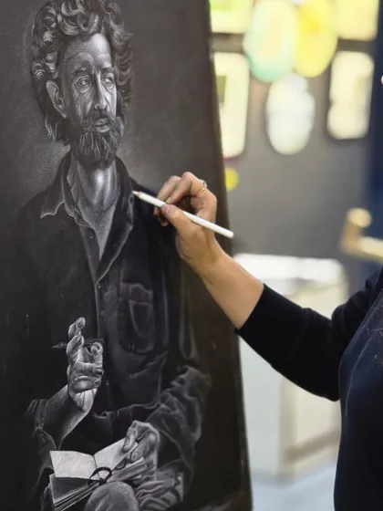 A student works on a realistic portrait of a man, using a glass marking pencil to capture the texture of his beard and the expression in his eyes.