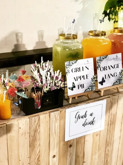 A rustic wooden bar setup just for kids. It features large glass dispensers of "Green Apple Pie" and "Orange Tang" juice, complete with fun straws and a welcoming "Grab a drink" sign.