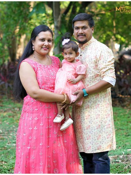 A classic family portrait taken outdoors. The family is dressed in coordinating pink and floral outfits, creating a cohesive and beautiful image against the natural green background.