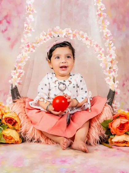 A happy baby girl sits in a floral swing, holding a red ball and smiling brightly. The pink floral theme is perfect for a sweet and cheerful portrait.