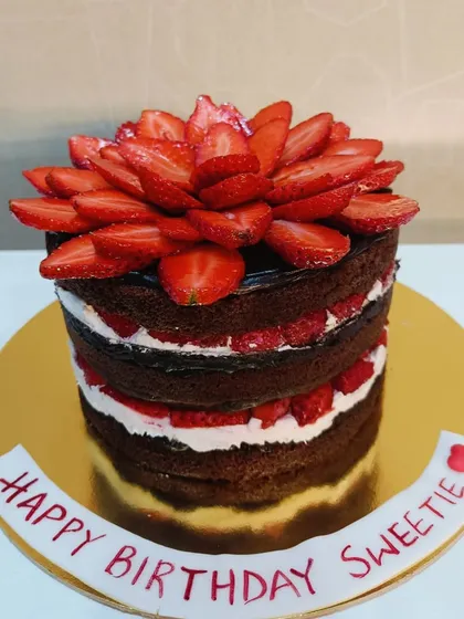 A naked chocolate cake with layers of fresh cream and sliced strawberries, topped with a beautiful strawberry flower.