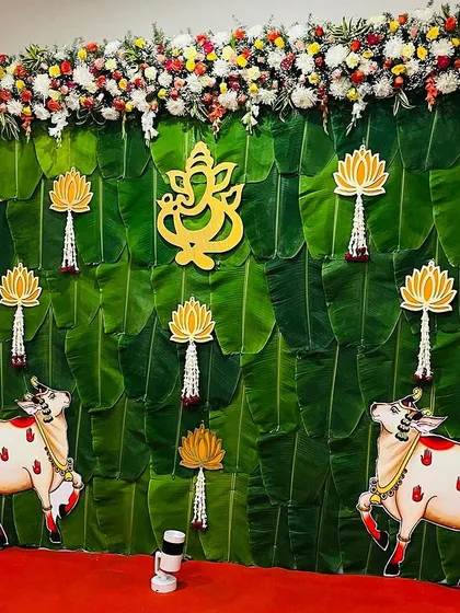 A full stage setup for a traditional baby shower. The backdrop is made of banana leaves with a Ganesha cutout, lotus hangings, and cow motifs.