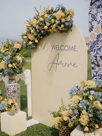 A chic and modern Haldi setup by the sea in Goa. The "Welcome, Amore" sign, paired with blue and yellow florals and patterned vases, creates a sophisticated coastal vibe inspired by Italian summers.