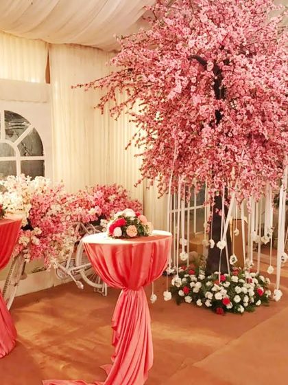 An indoor event space decorated with a large artificial cherry blossom tree and pink-draped cocktail tables, creating a whimsical, garden-like atmosphere.