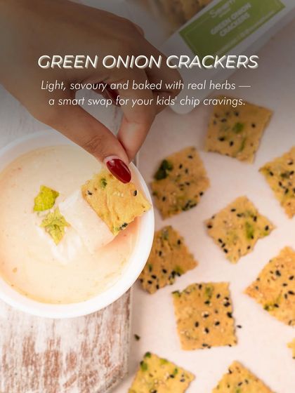 A hand dipping one of my Green Onion Crackers into a creamy dip. Light, savoury, and baked with real herbs, they are a smart and delicious swap for your kids' chip cravings.